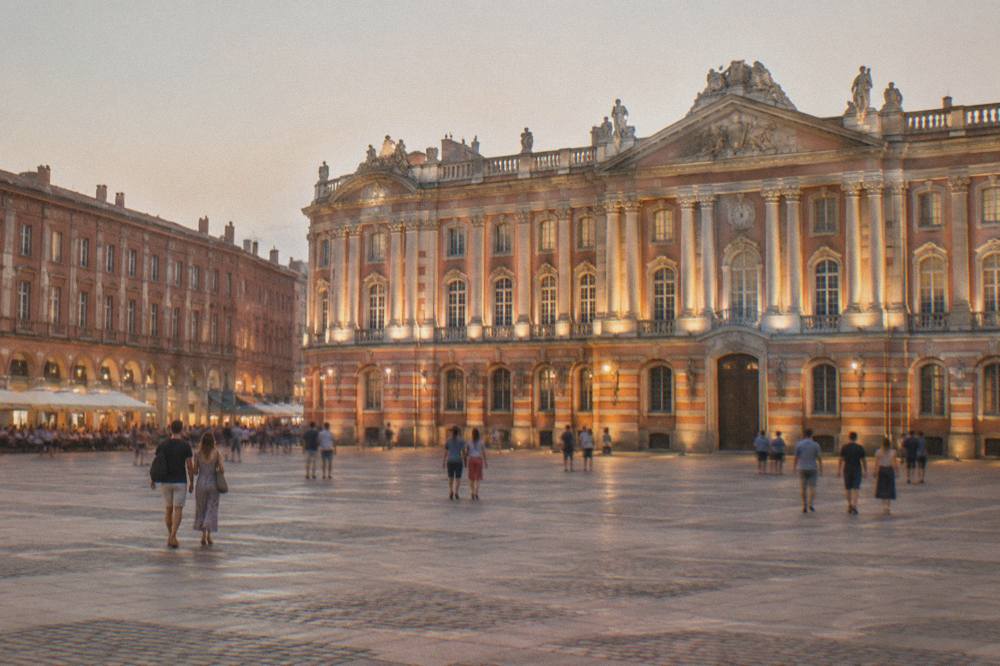 The illuminated front of the Capitole building in Toulouse at night with people walking across the square