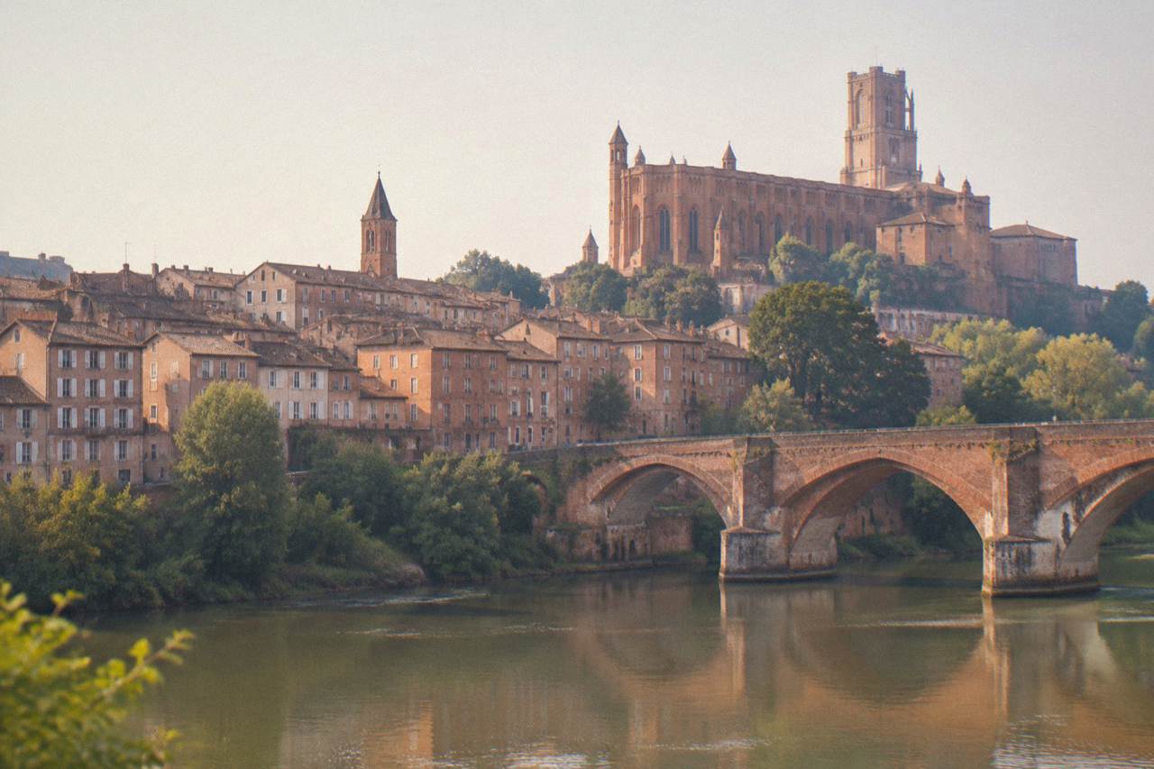 A small French town of warm-toned brick buildings seen from across a river, with a church tower rising above the rooftops