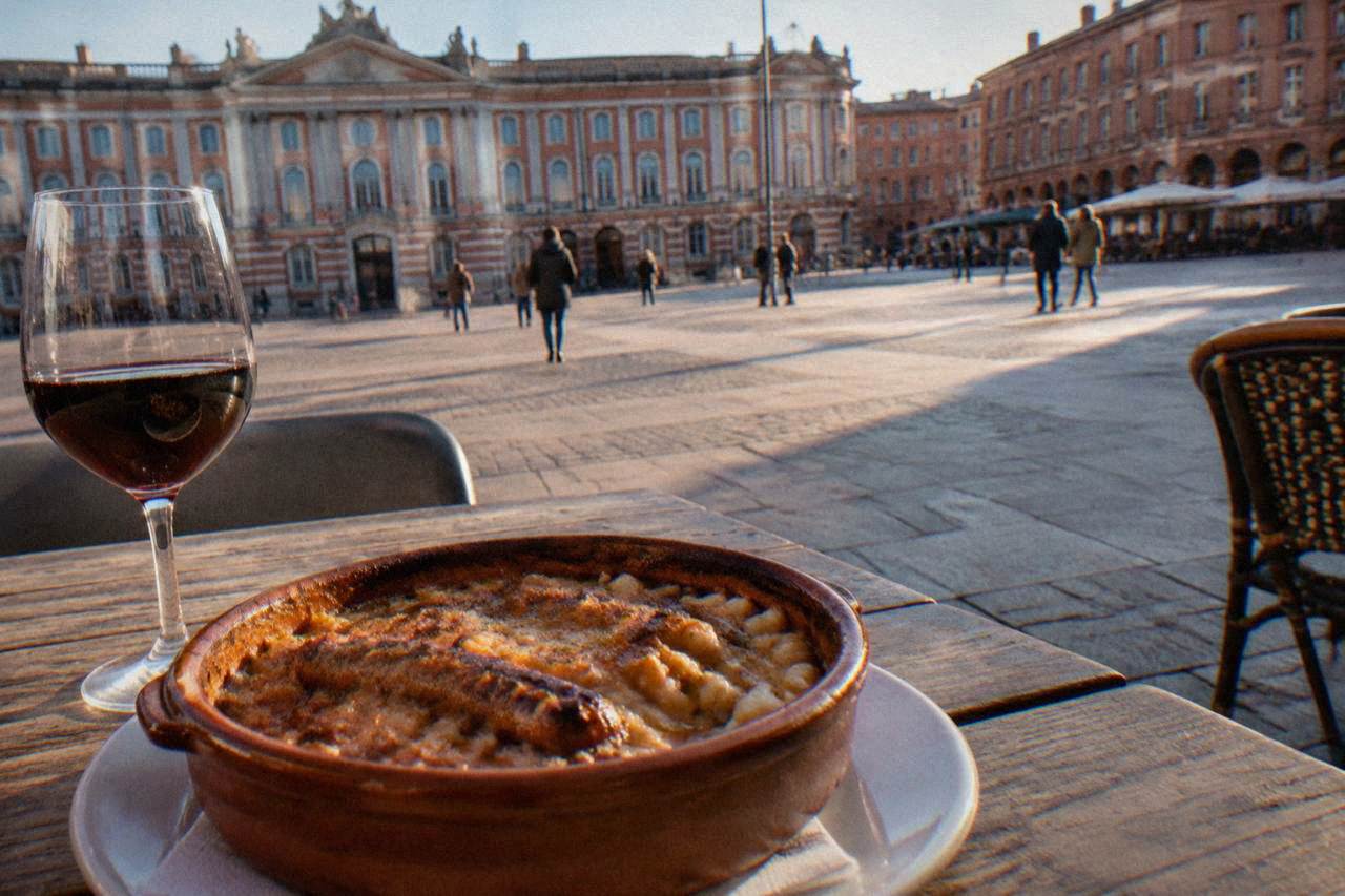 A clay cassoulet dish with a golden crust beside a glass of red wine on a wooden table