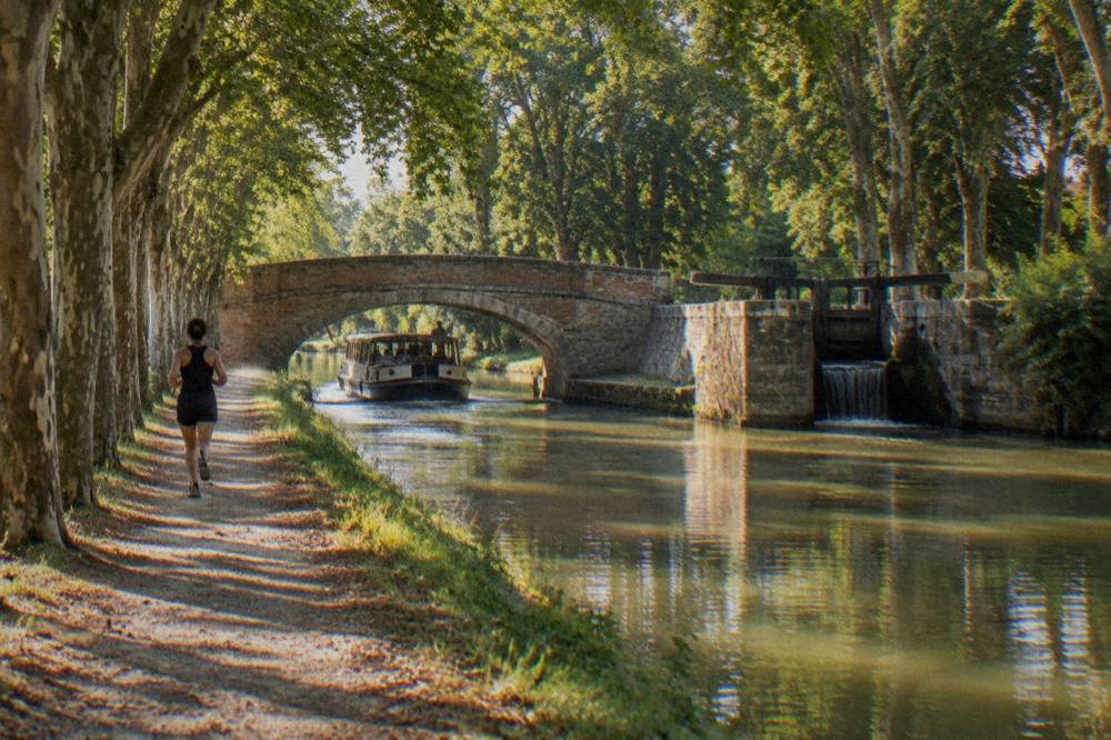 A flat canal boat passing under a stone bridge on the Canal du Midi