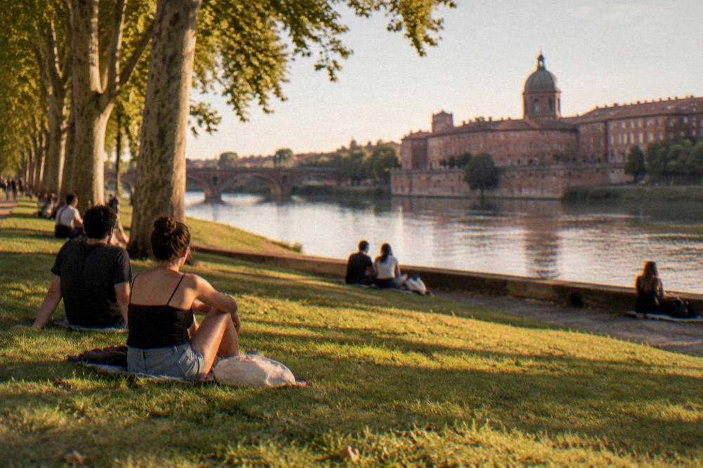 Visitors sitting on the grass of a riverside lawn beside the Garonne on a sunny day