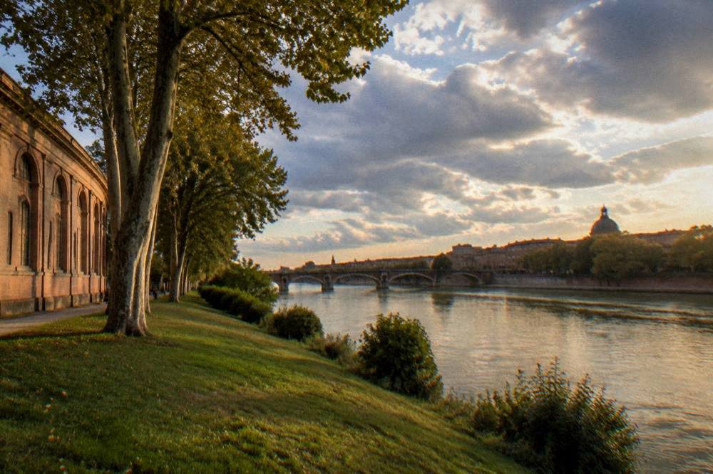 A wide view of the Garonne river at dusk with a bridge and city rooftops beyond