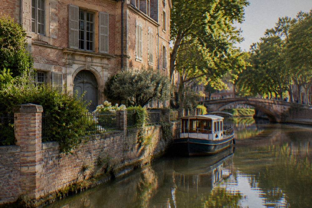 A tree-lined canal with a moored boat and a stone bridge reflected in still water