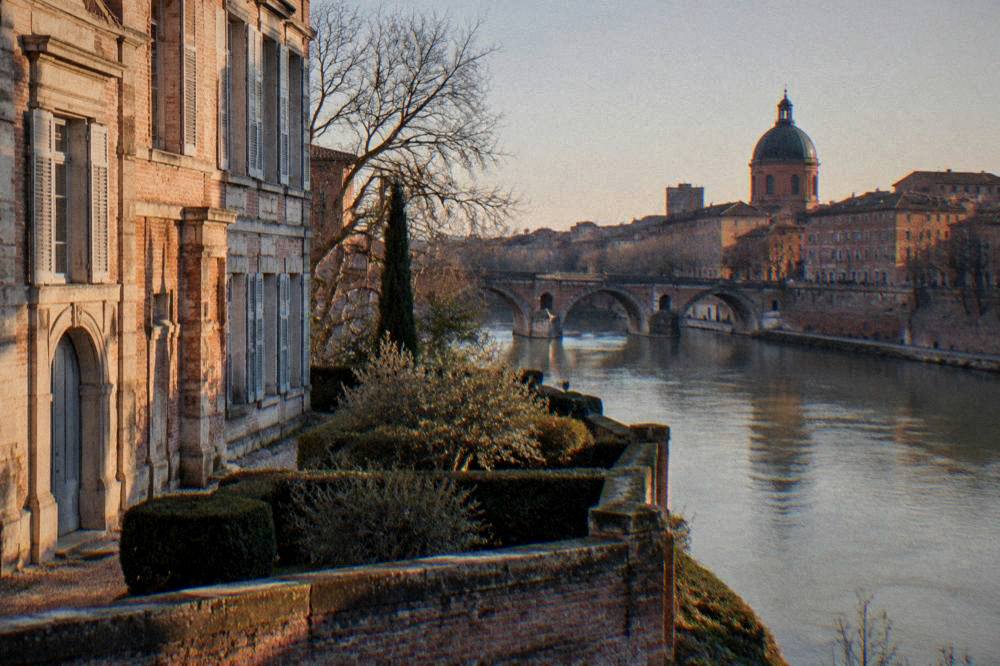 An evening view of a riverside city with warm light on old brick buildings and a stone bridge