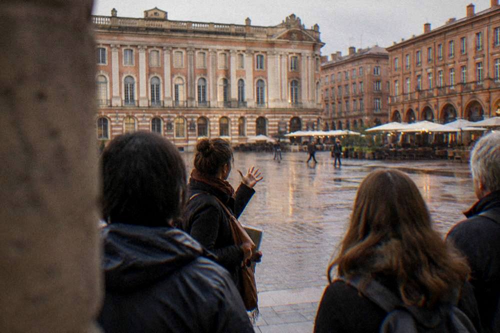 A guide speaking to a small group of visitors on a Toulouse square lined with pink brick buildings