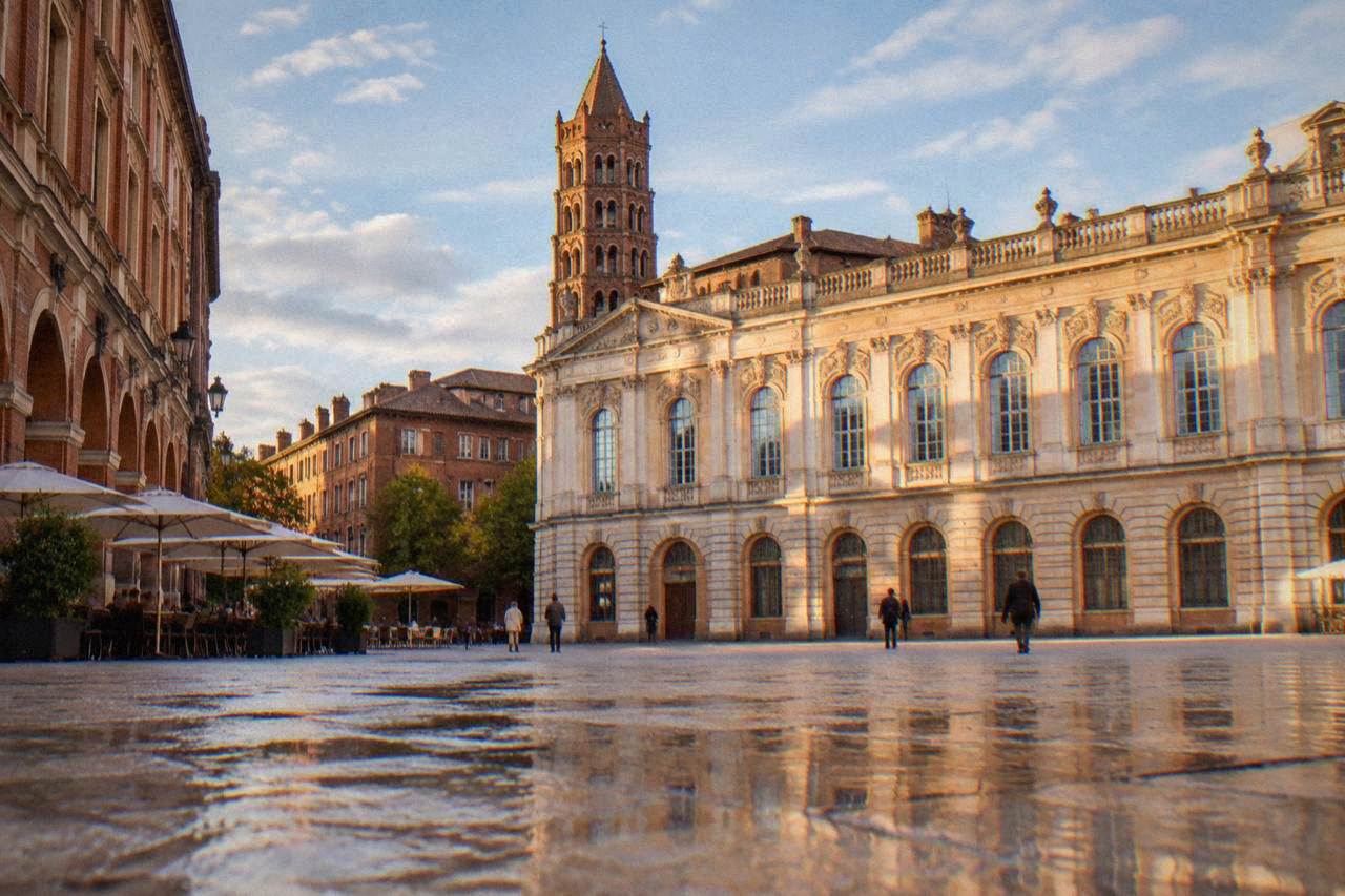 A pale stone museum facade on a square in Toulouse with a brick basilica tower rising behind it