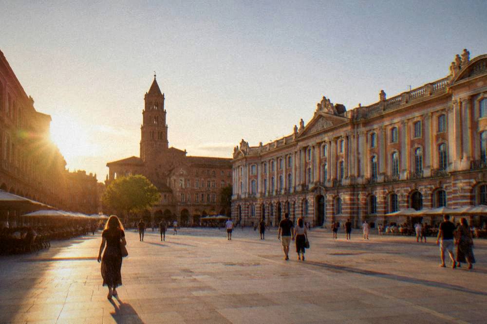 A wide square in Toulouse at golden hour with a brick church tower and people walking