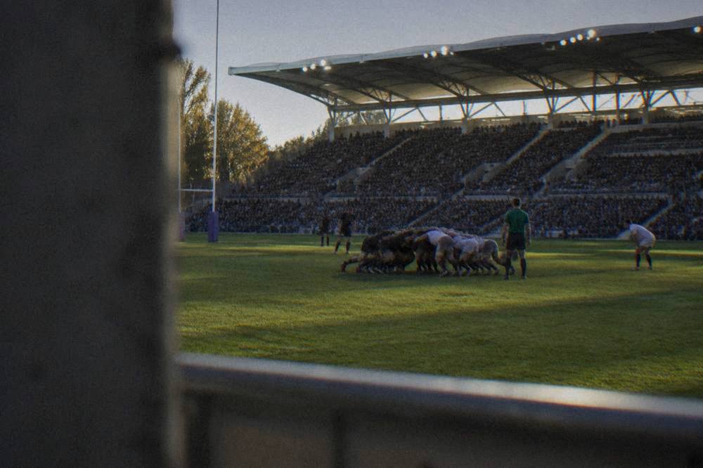 Rugby players in a scrum on a green pitch under floodlights, with packed stands behind them