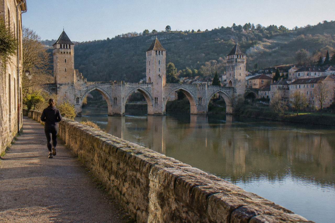 A medieval fortified bridge with three stone towers crossing a calm river through a green valley town