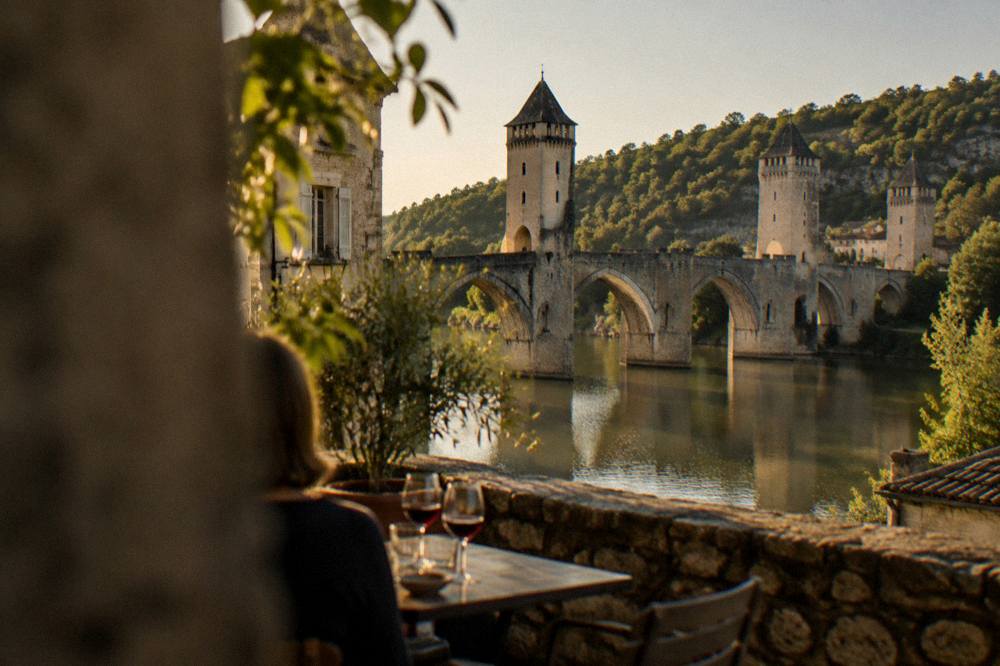 A shaded terrace table with glasses of red wine beside a stone wall in an old town square