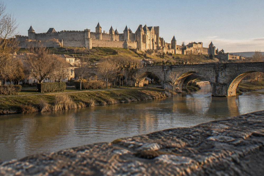 Fortified city walls and towers seen across a river bridge in warm evening light