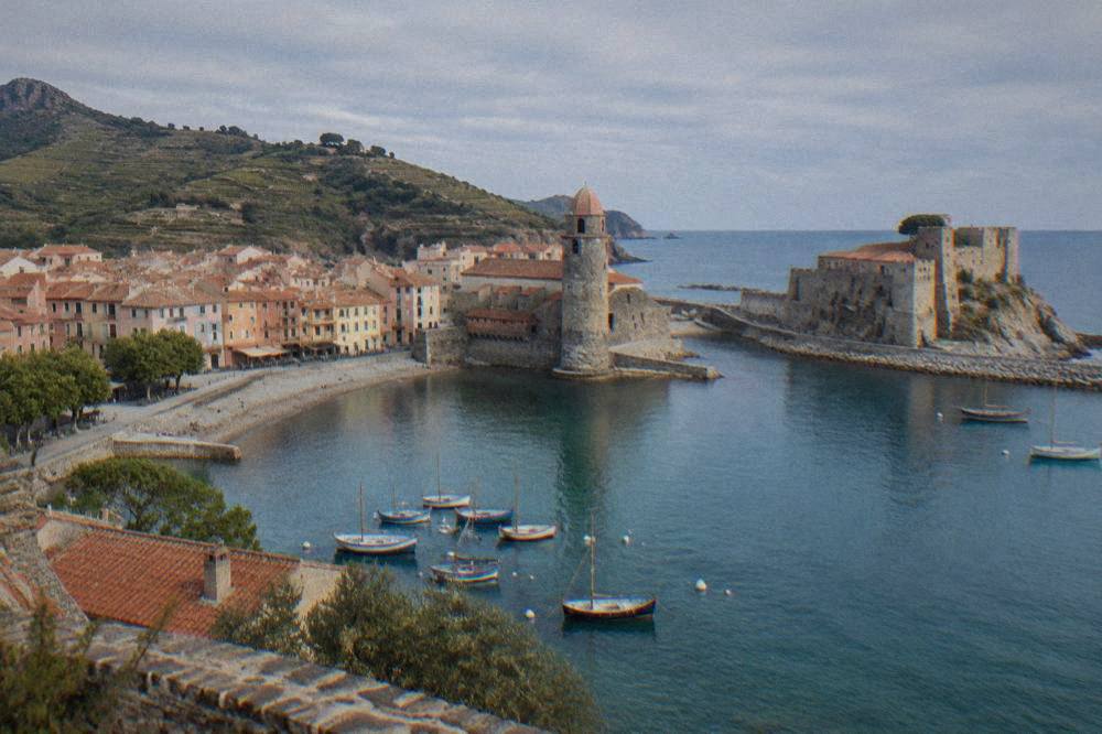 A stone castle standing at the entrance to a small Mediterranean harbor with boats