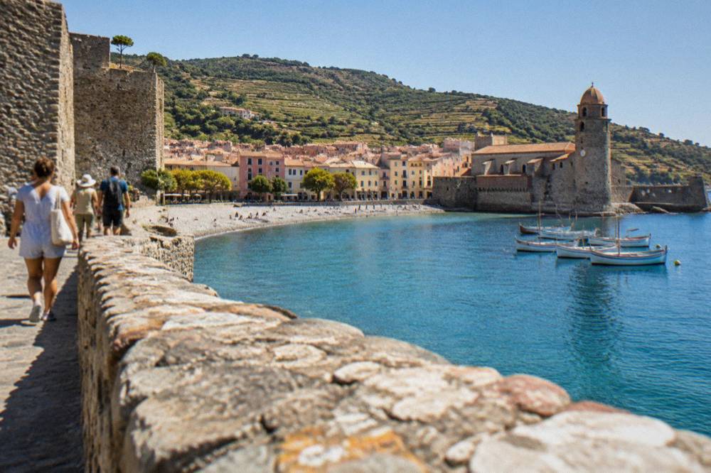 Tourists walking beside a stone castle wall above a Mediterranean bay