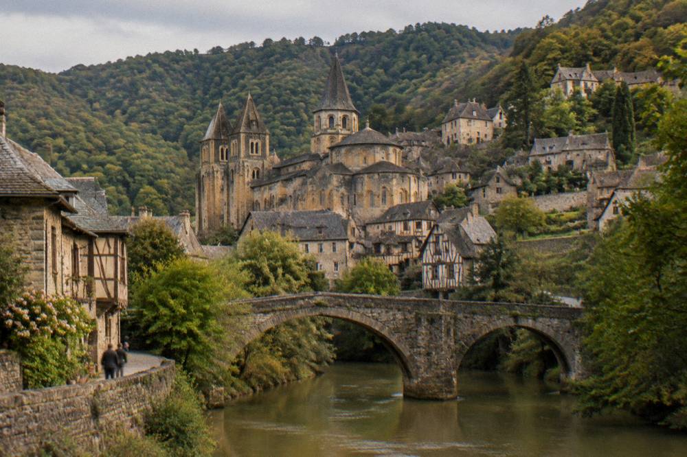 Stone bridge over the Dourdou river with the village of Conques rising on the wooded hillside beyond