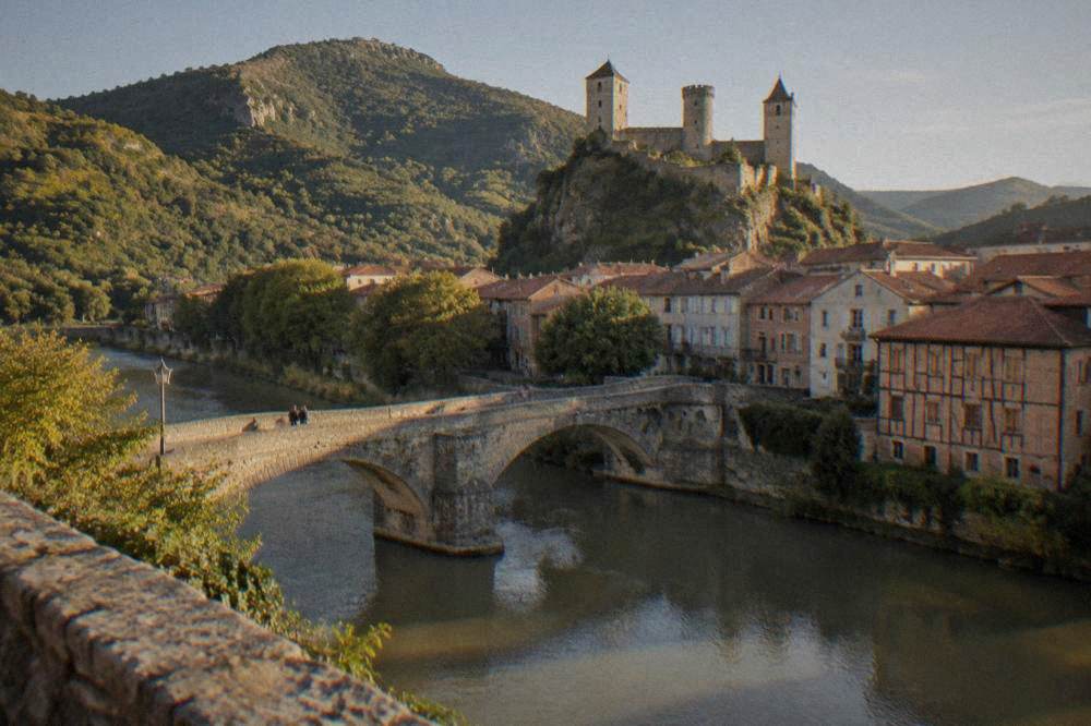 A pedestrian stone bridge over a clear river with a castle visible in the distance