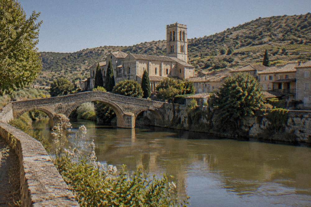 An old stone abbey church with a bell tower seen across a river from a village bank