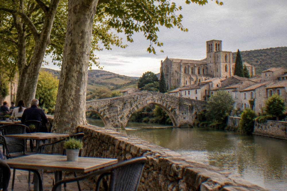 A stone terrace with tables under plane trees beside a river in a French village