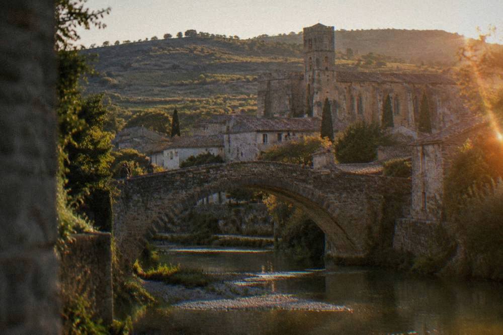 A stone arch bridge over a shallow river with a village and church tower behind it at golden hour