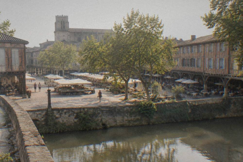 A wide medieval square with stone and timber arcades and cafe tables under the walkways