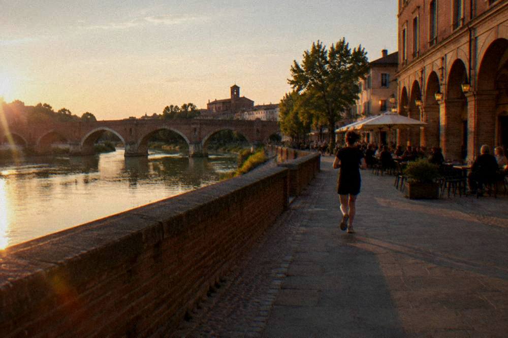 An arcaded square at dusk with warm lamplight on pink brick facades and people at outdoor tables