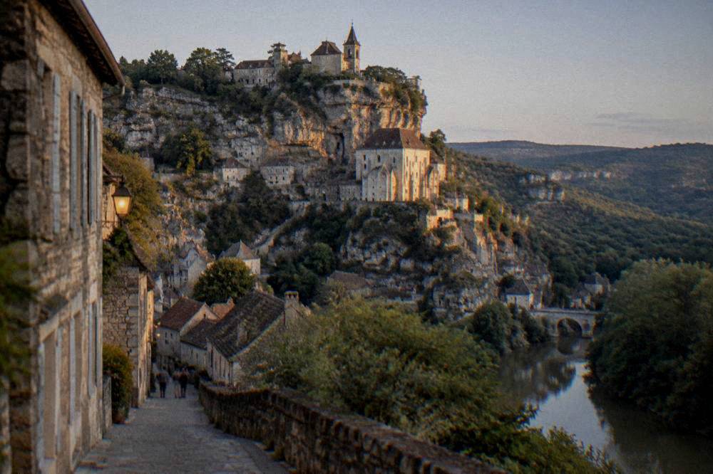 A village street in warm evening light with stone facades and a view of hills in the distance.