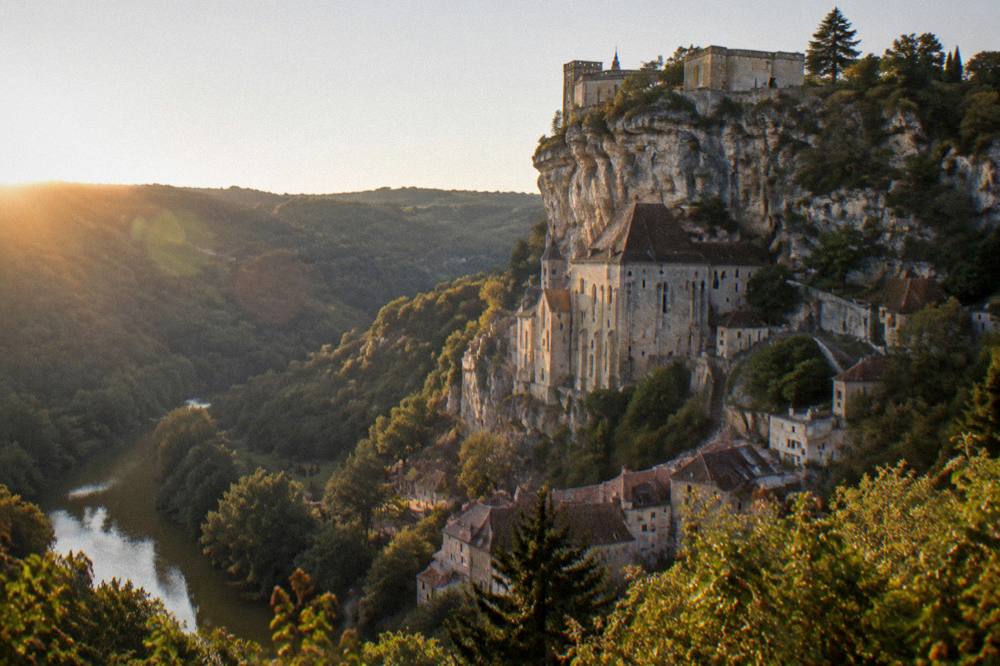 A stone castle wall at the top of a cliff overlooking a wooded valley in the late afternoon.