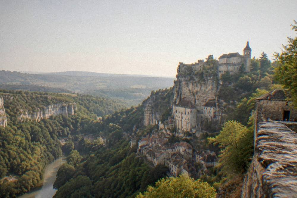 A view from a cliff-top castle wall across a wooded gorge toward hills fading into haze.