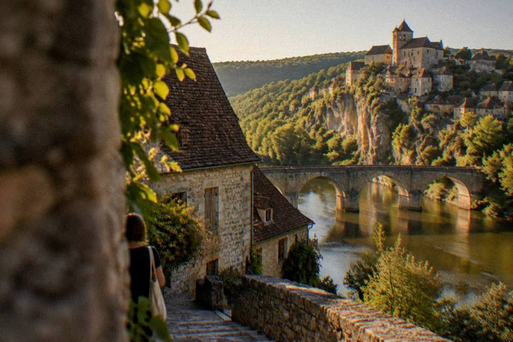 A stone bridge over a calm river with a clifftop village in the background at golden hour