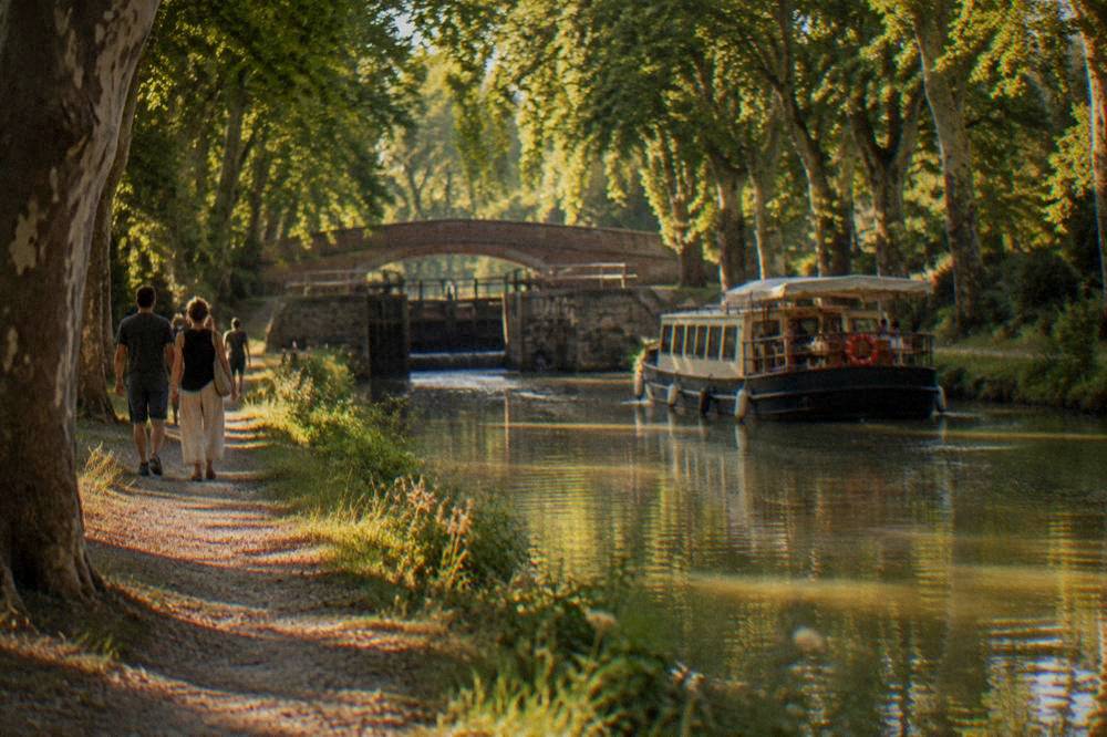 A tree-shaded canal towpath with walkers and a slow boat
