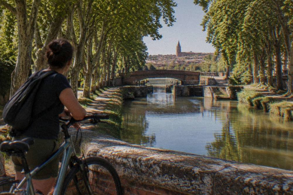 A cyclist stopped on a canal bridge looking back toward pink brick city rooftops in the distance