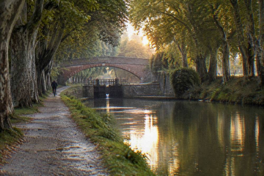 An evening canal scene with warm light on the water, a towpath cyclist in the distance and a stone bridge