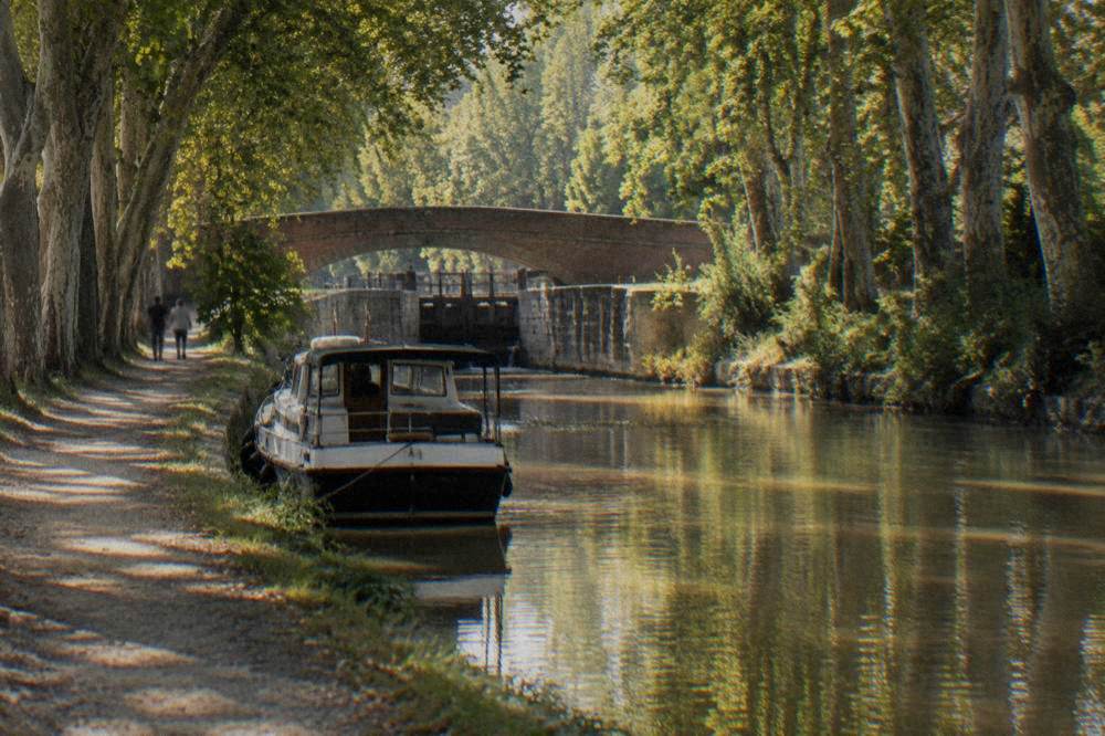 A tree-lined canal with a small boat moored beside a stone towpath