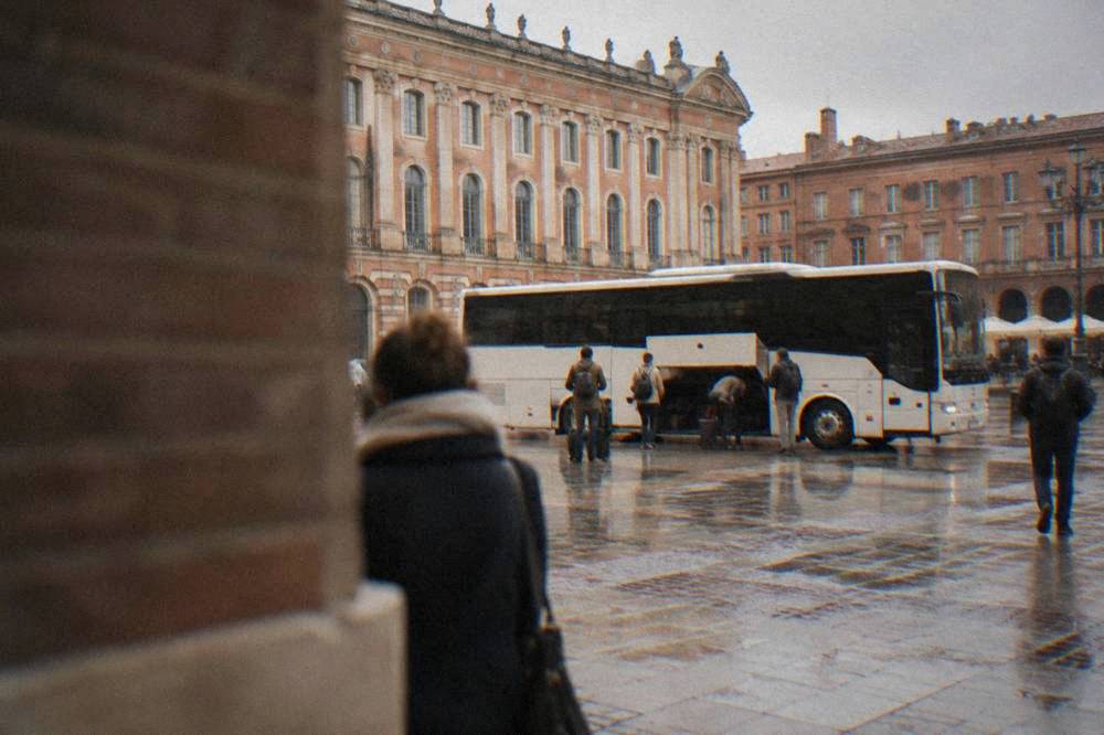 A white airport shuttle bus parked at a curb outside an airport terminal with passengers loading luggage