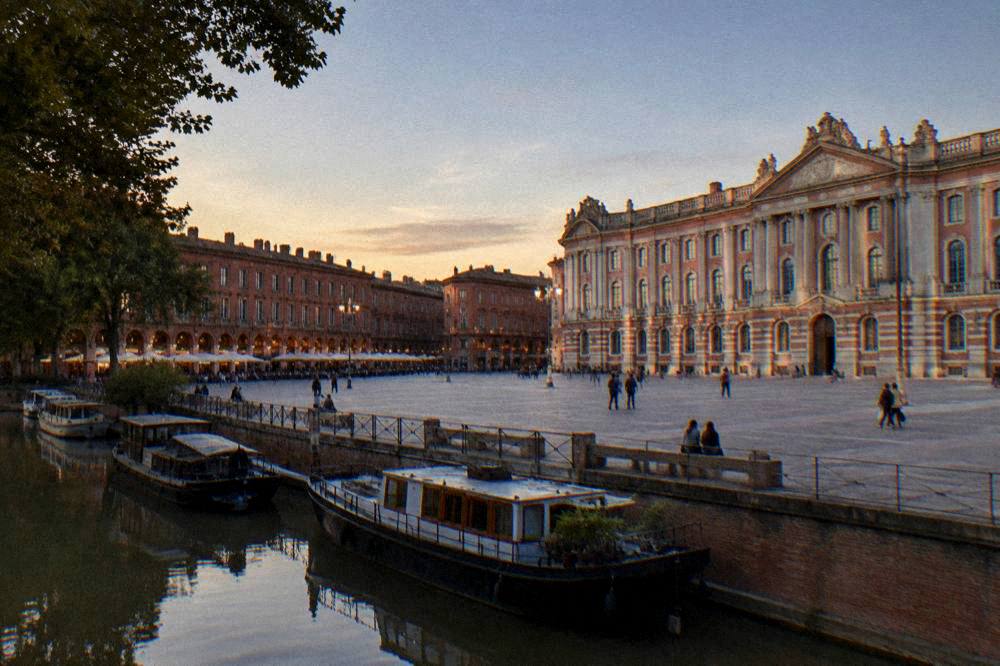 A tree-lined canal with moored boats passing brick warehouses in Toulouse