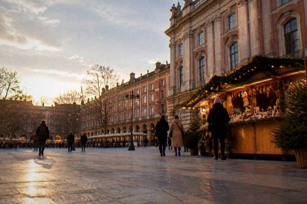 A lit Christmas market stall in front of a pink brick building on a Toulouse square at dusk