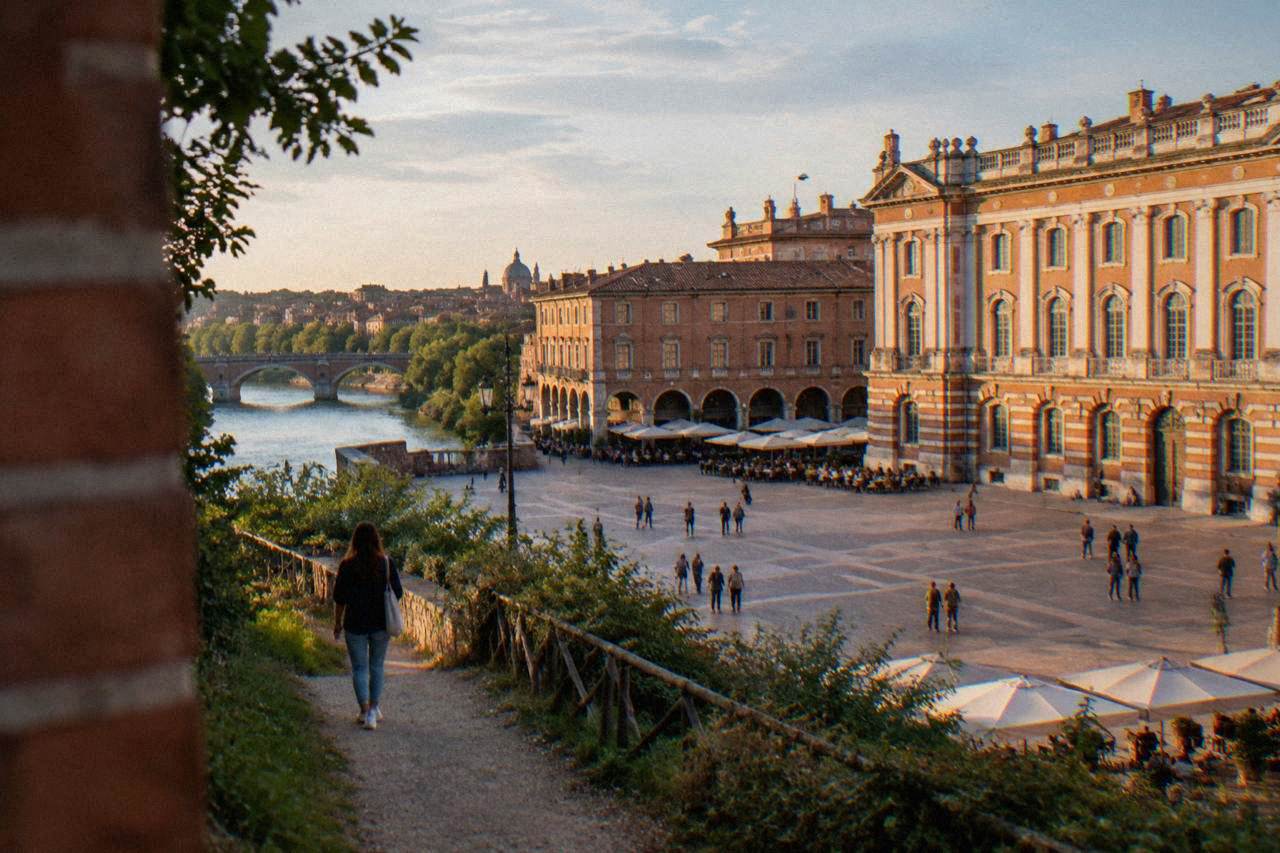 A walker on a dirt path with the pink brick rooftops of Toulouse and the Garonne river in the background