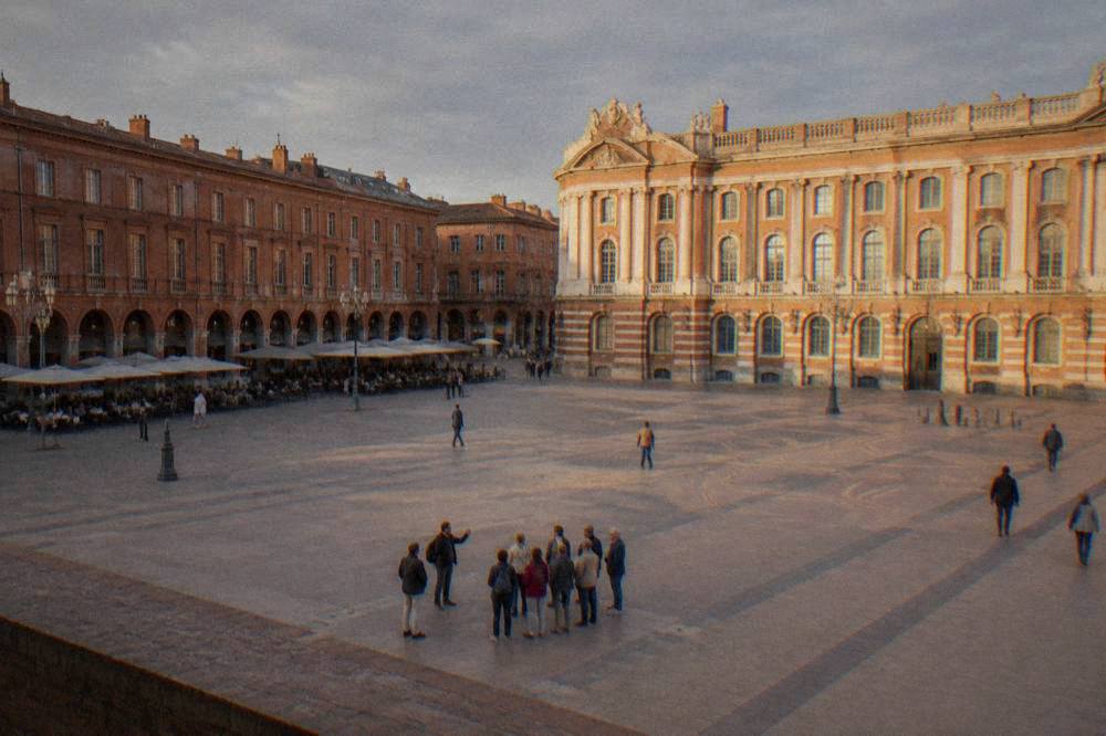 A small guided walking group pausing in a sunlit Toulouse square with pink brick facades