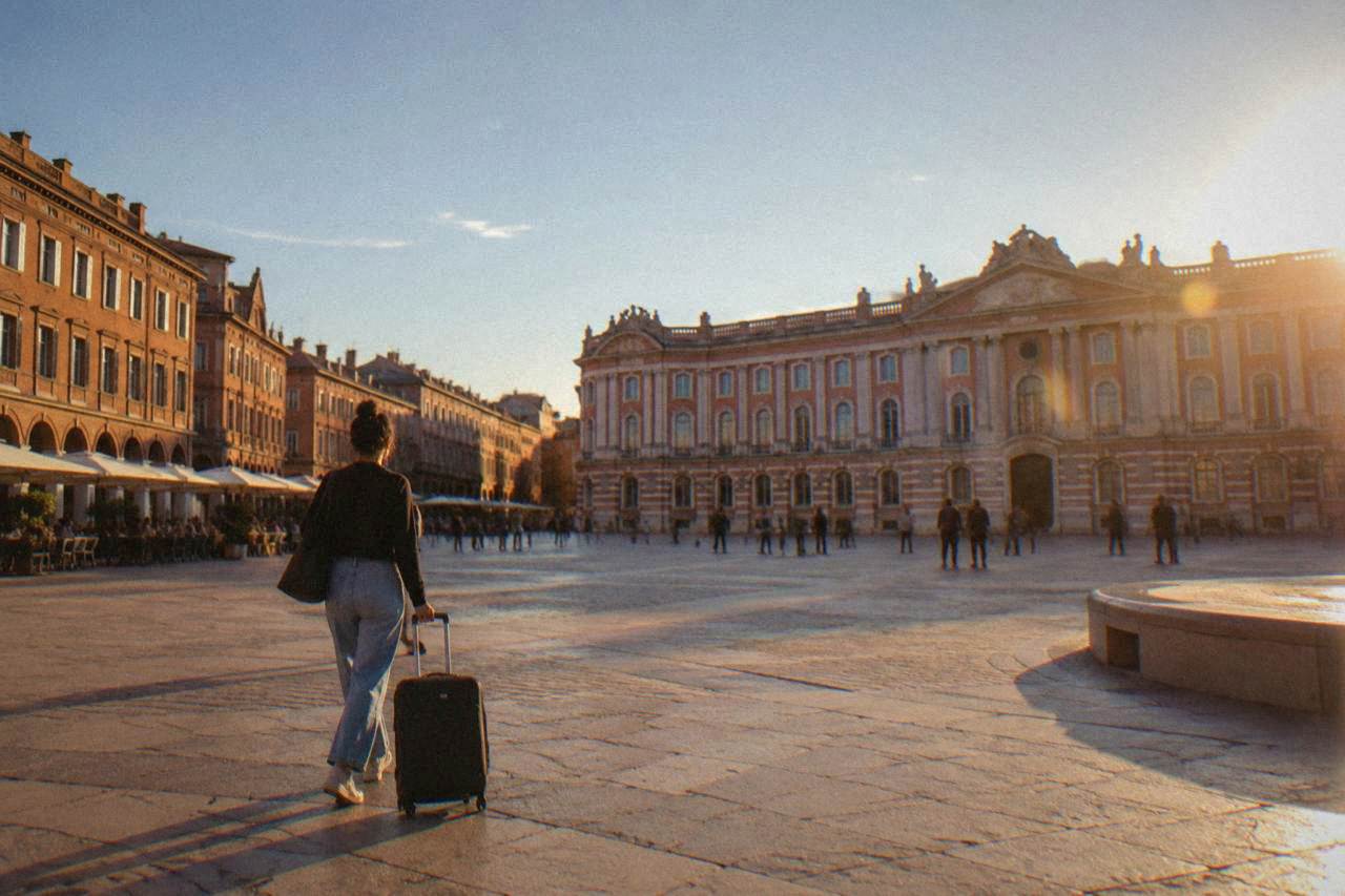Traveler with a rolling suitcase walking past pink brick buildings on a sunny Toulouse street