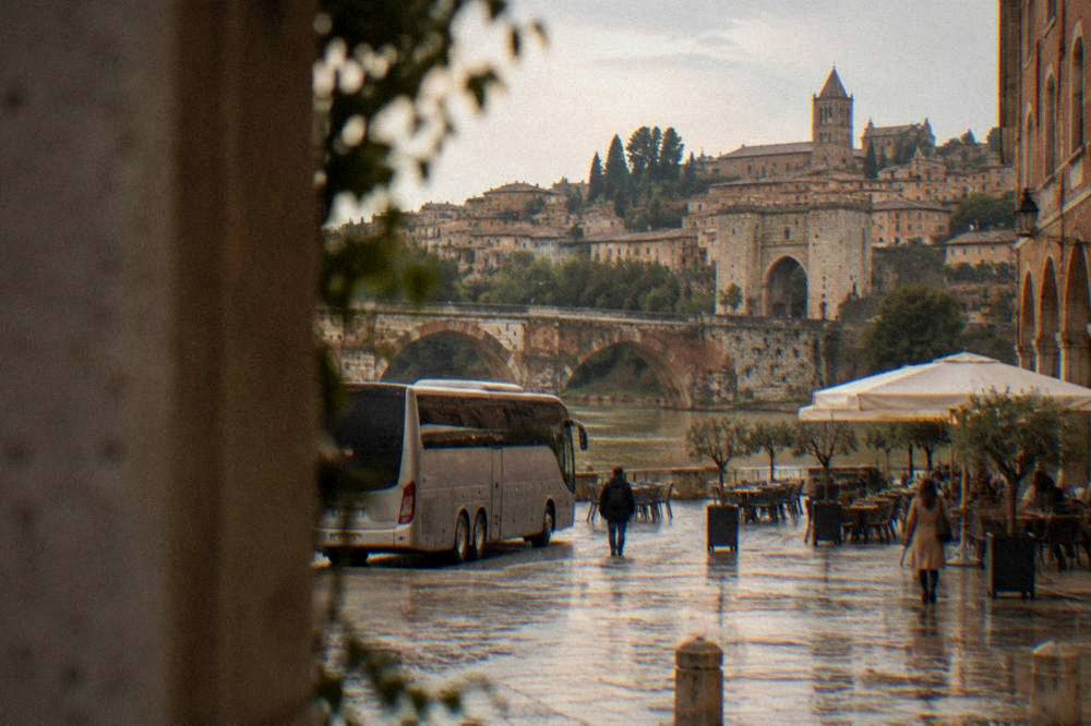 A coach parked outside a medieval hilltop town with a stone archway and small cafes