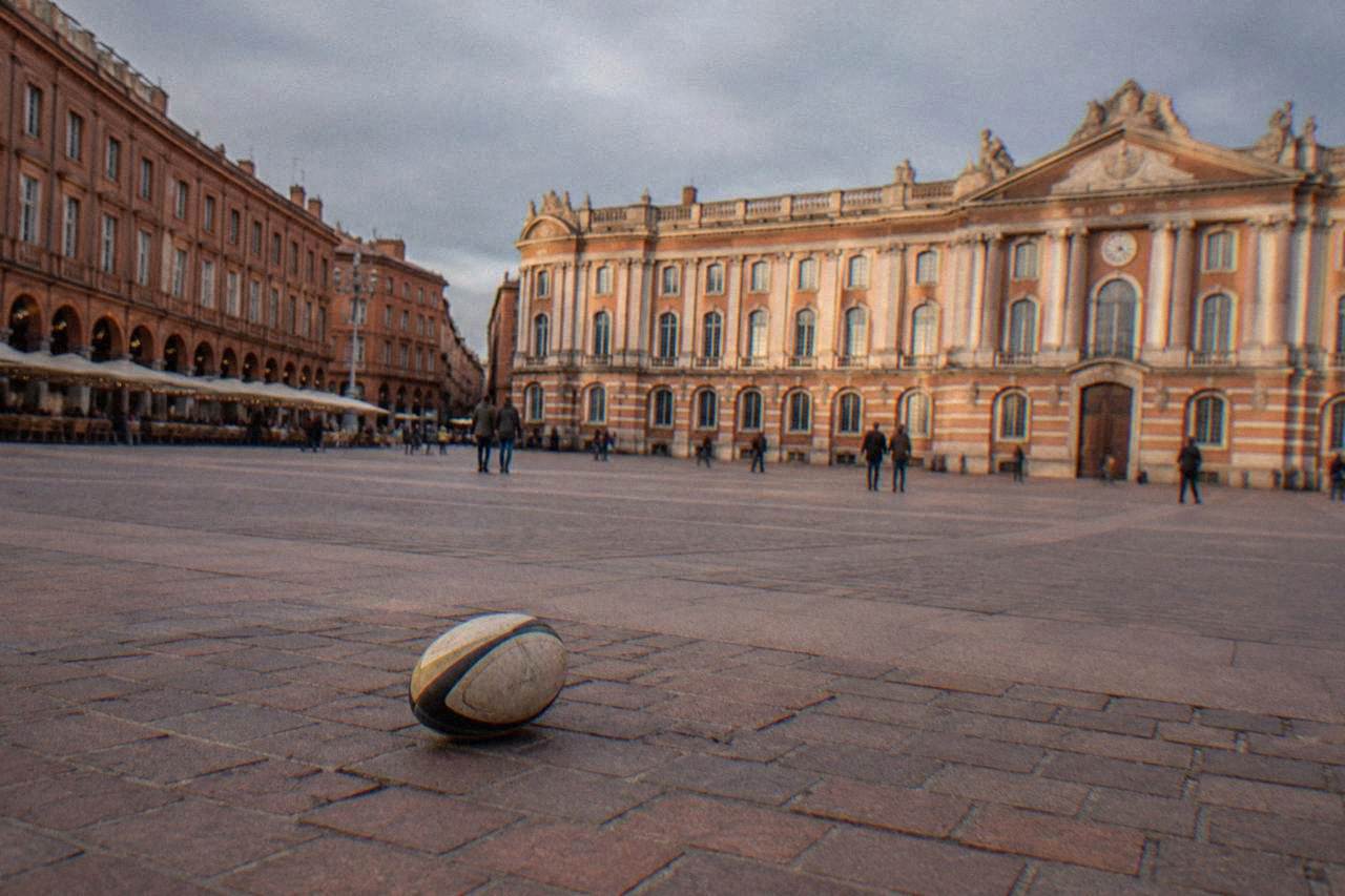 A rugby ball resting on a pink brick plaza in Toulouse with the Capitole building in the background