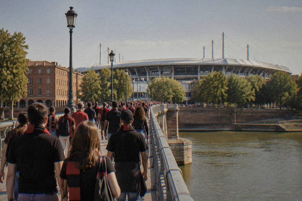 Fans in red and black scarves walking across a bridge over the Garonne toward a stadium
