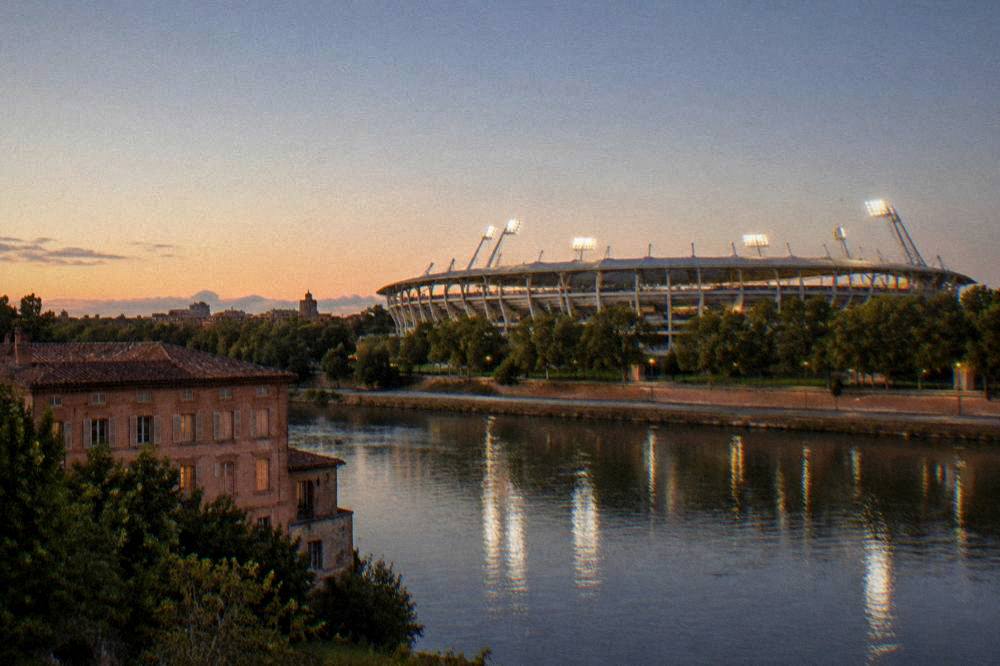 A evening view of a floodlit stadium from across a river with pink brick buildings in the foreground