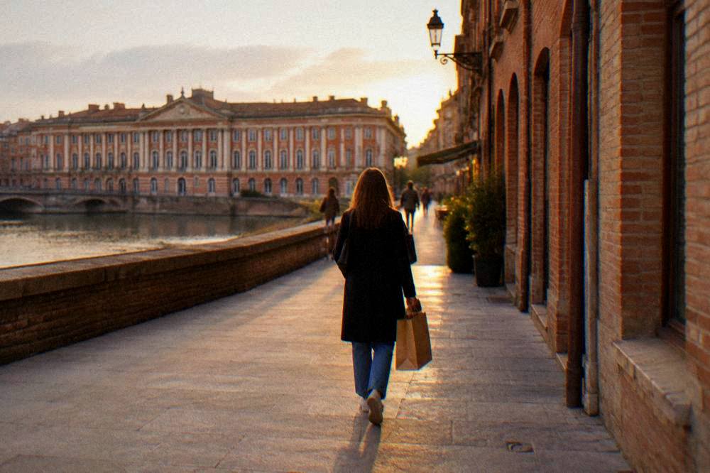 A shopper carrying a paper bag past pink-brick facades in a Toulouse side street at golden hour