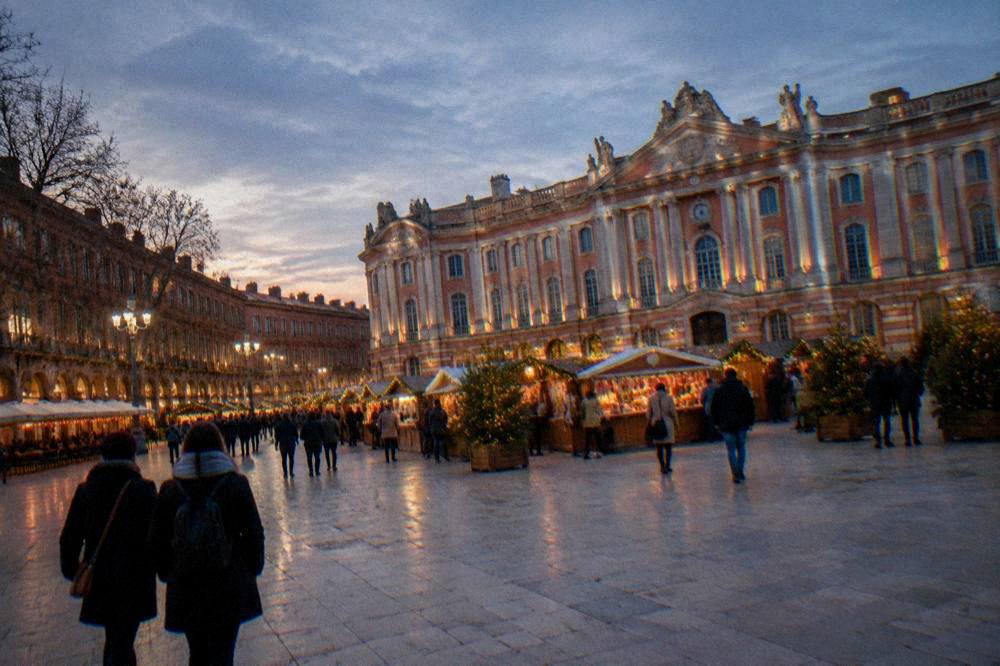 Festively lit Christmas market stalls in front of a grand pink brick city hall