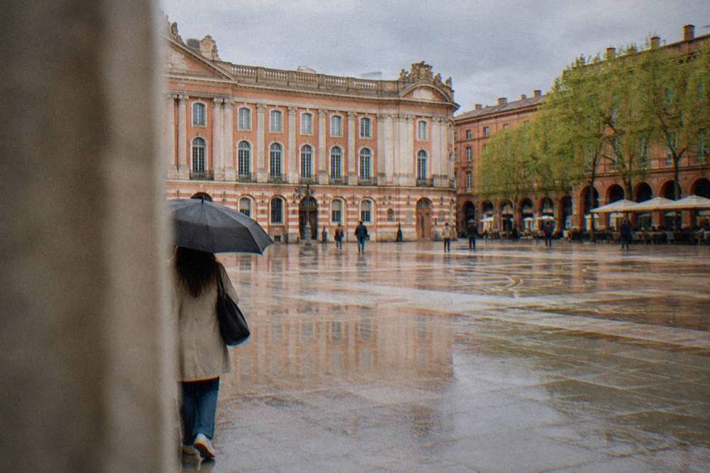 A visitor with an umbrella walking past pink brick buildings on a showery spring day in Toulouse