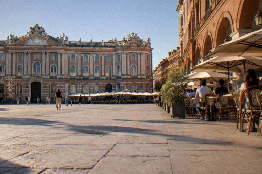 A sunny Toulouse square with cafe tables and pink brick arcades around the edges