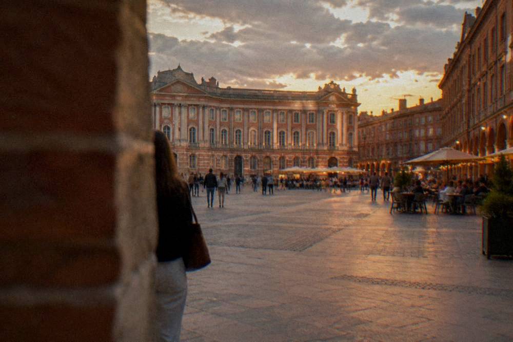 A city square at dusk with lit cafe terraces and pink brick facades